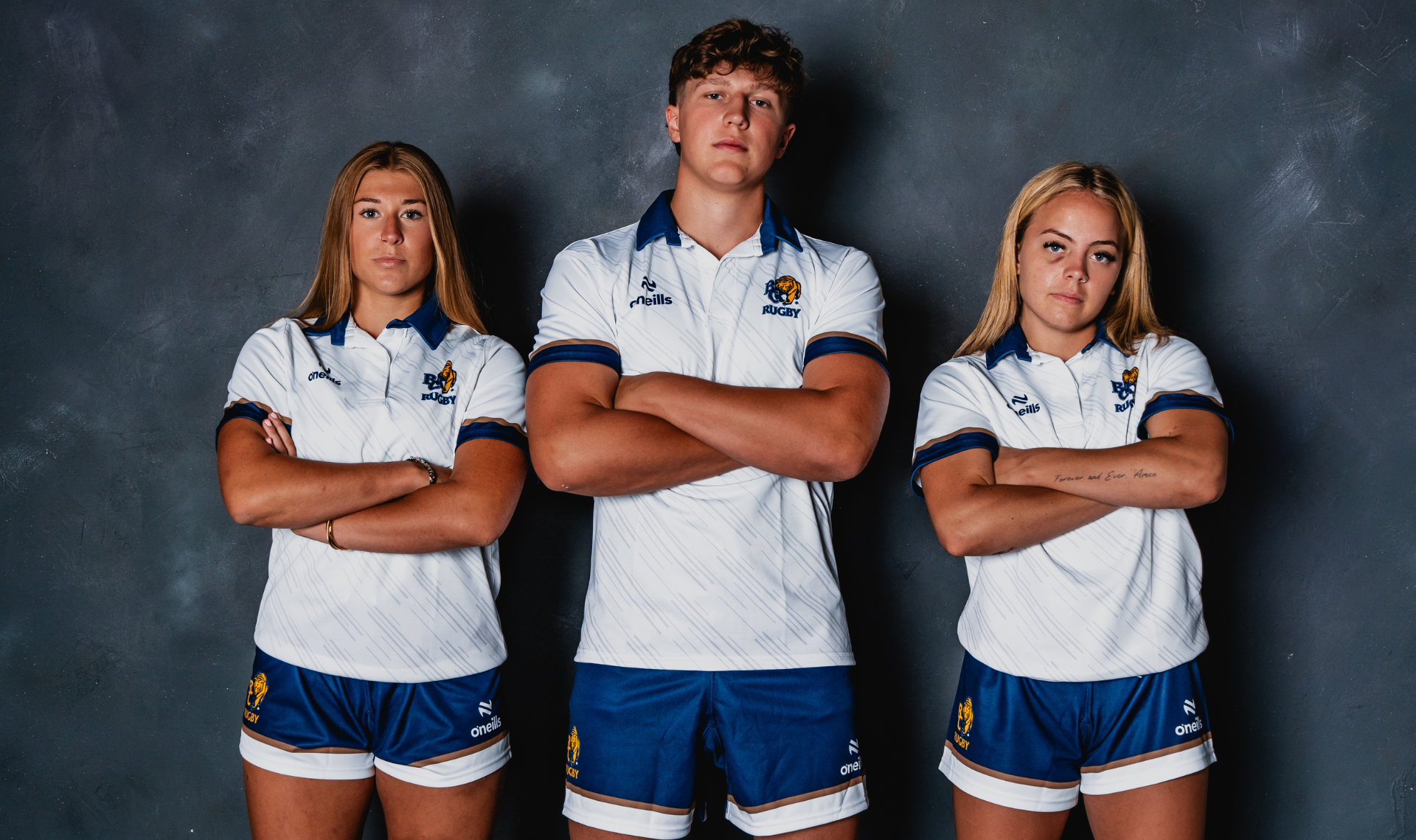 Three athletes in white-and-blue uniforms stand with arms crossed against a dark backdrop.