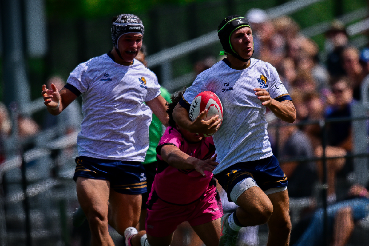 Two rugby players in white shirts sprint with a ball during a match on a grassy field