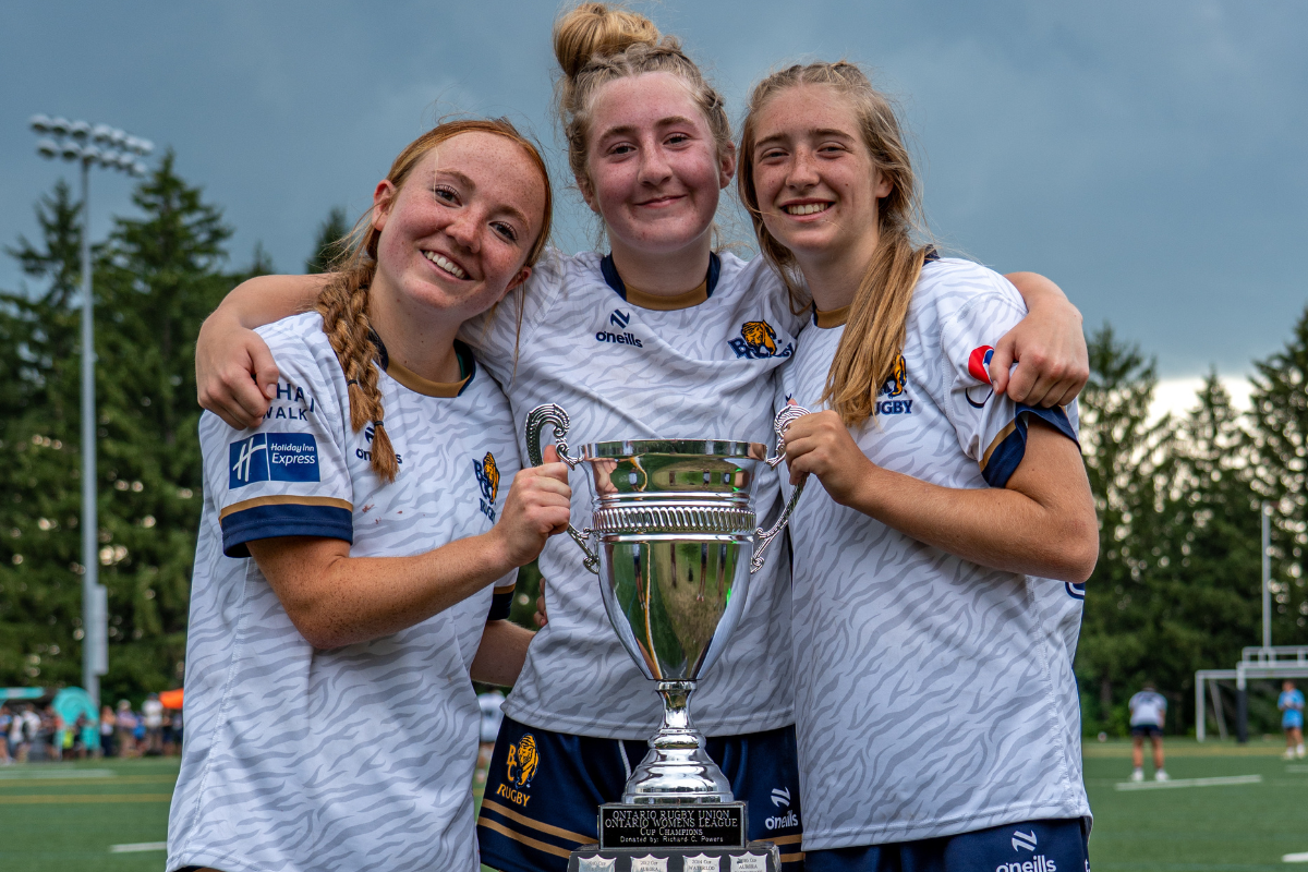 Three soccer players in white jerseys smile while holding a trophy on a field.