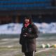 Person in a dark coat and red hat standing on a snowy field at dusk, with blurred buildings in the background