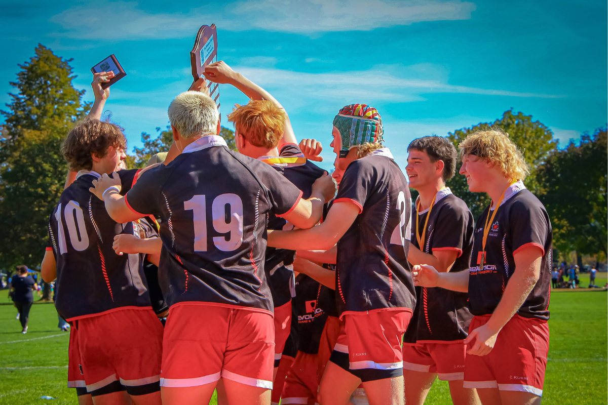Soccer players in black and red uniforms celebrate on a sunny field, arms raised in a team huddle