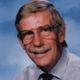 Portrait of a smiling man with glasses and a mustache, wearing a suit and tie against a blue background