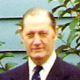 Portrait of a man in a dark suit and tie standing outdoors with greenery behind him