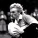 A man in a dark jacket leans over while hugging a rugby ball in a black-and-white photo.