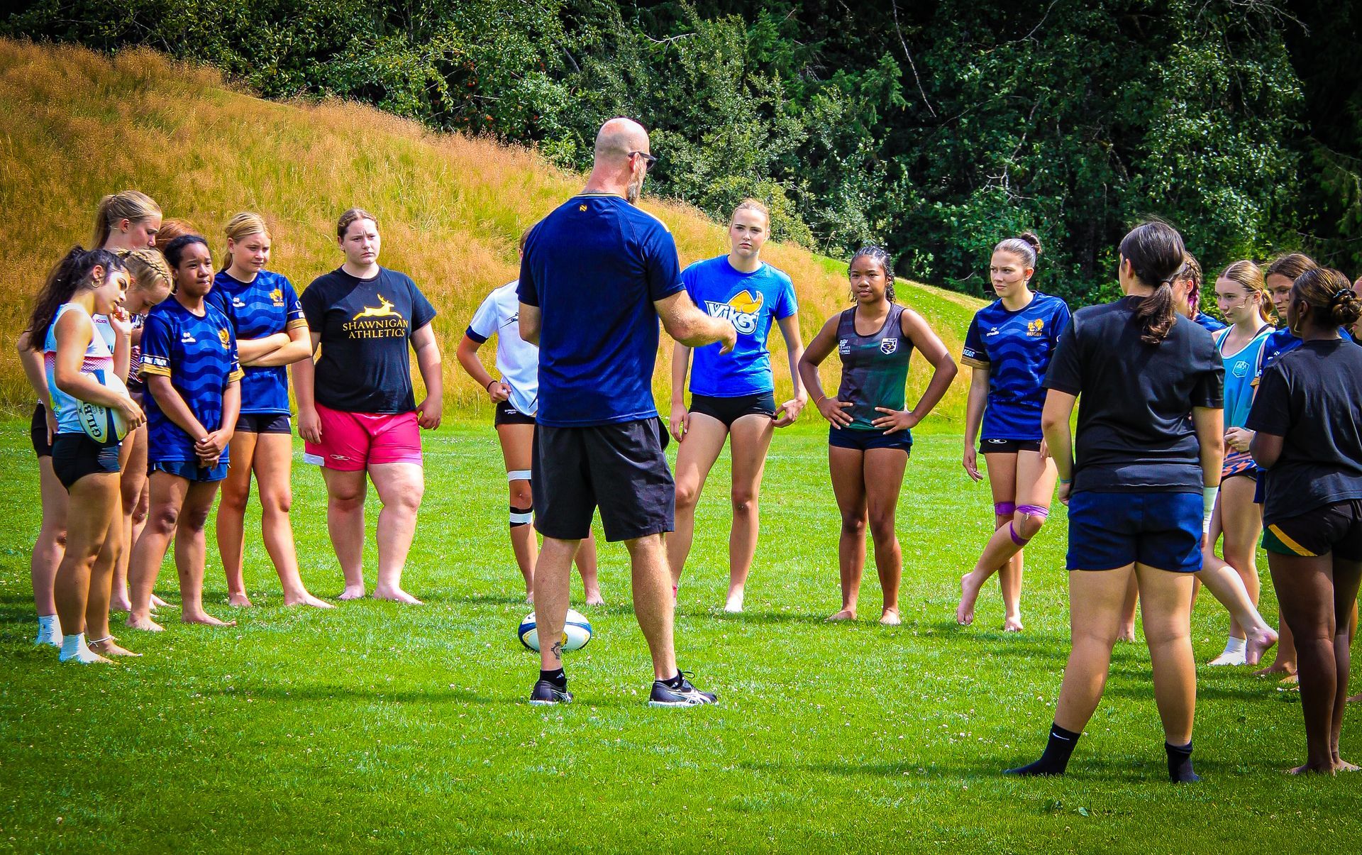 Coach briefing a group of athletes in blue shirts standing on a grassy field outdoors.