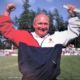 Man smiling with arms raised on a sports field, wearing a red, white, and navy jacket.