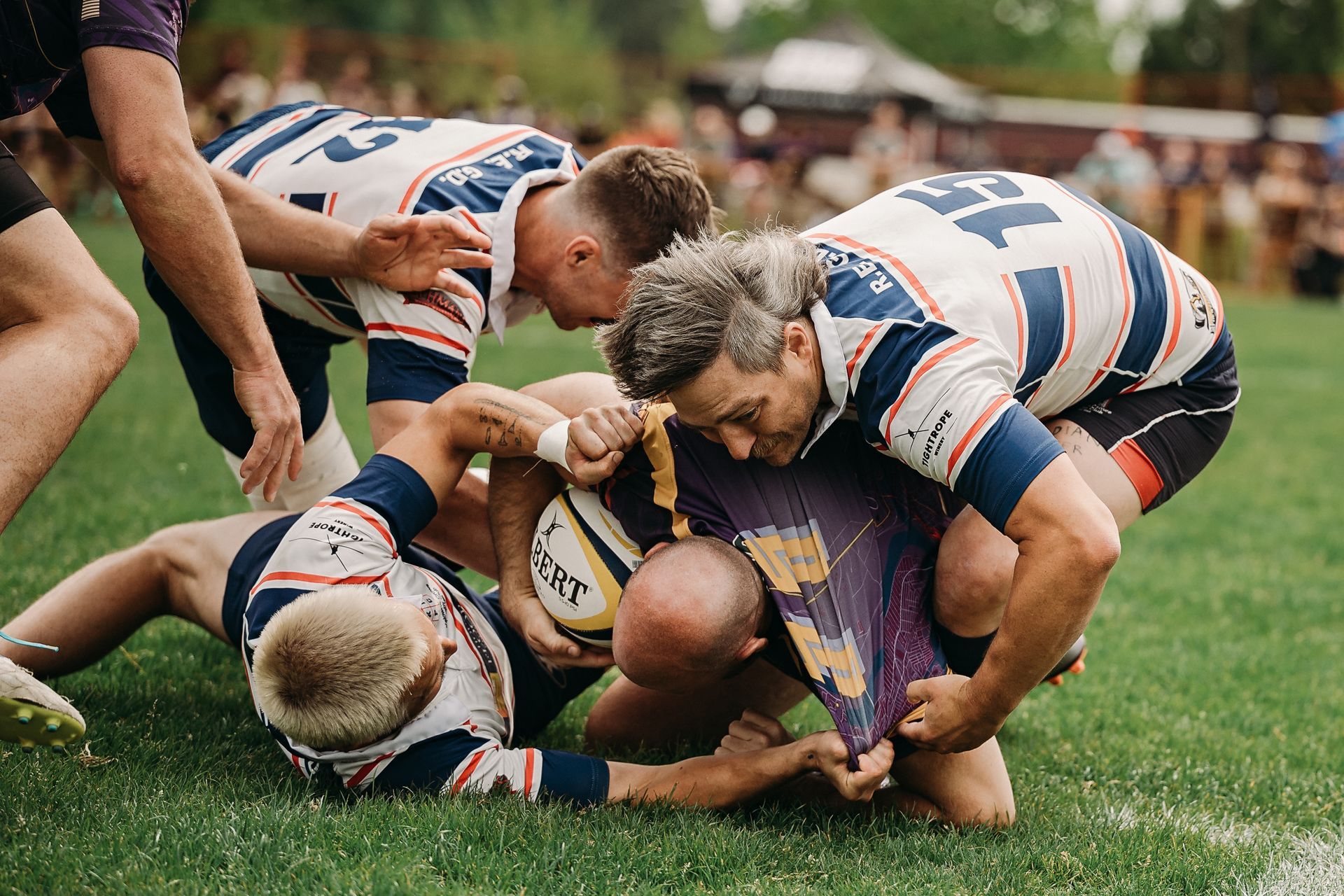 Rugby players in white and blue uniforms tackle an opponent holding the ball on a grassy field.