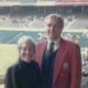 Two people standing together in a stadium, one in a red jacket and both smiling.