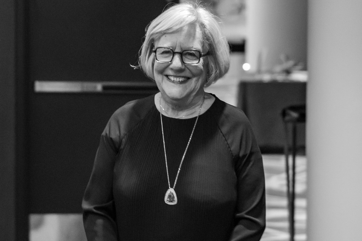 Smiling person with glasses and a pendant necklace, standing indoors against a blurred background.