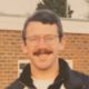 Smiling man with glasses and mustache standing outdoors in a collared shirt.
