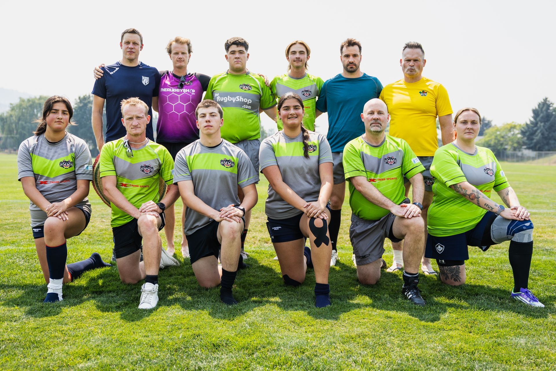 Rugby team posing on grass, with players in green and gray jerseys and coaches standing behind them.
