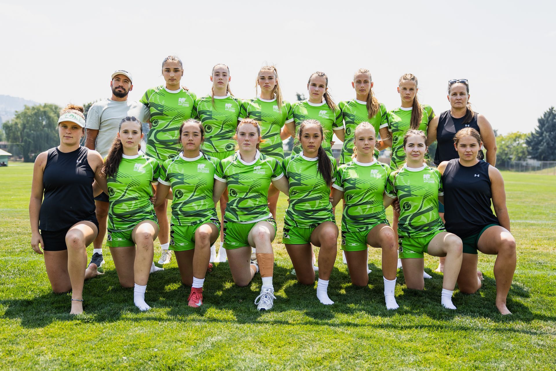 Twelve athletes in green uniforms posing outdoors on grass, with two people seated at the front.