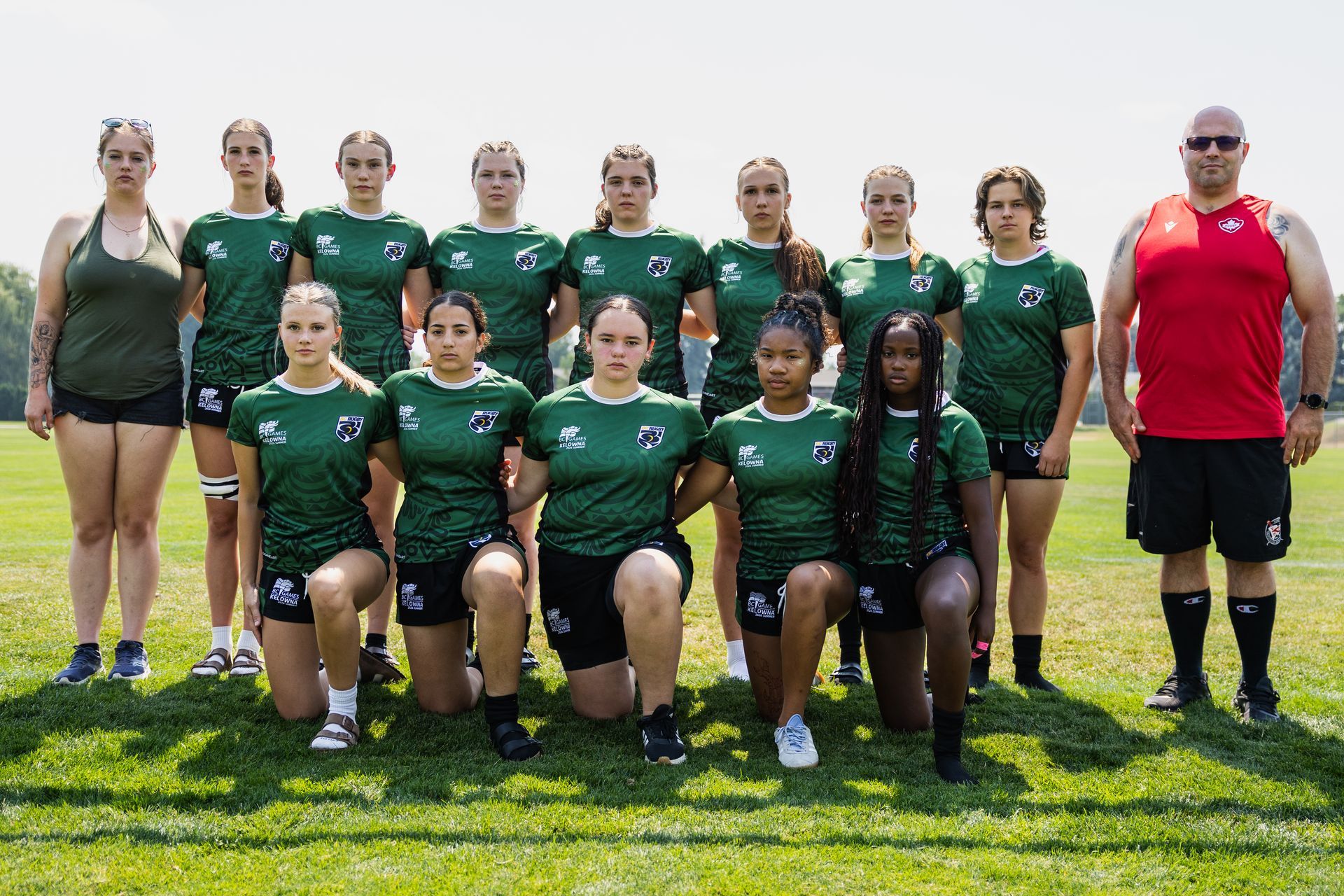 Group of athletes in green jerseys posing on a grassy field with two coaches at the ends.