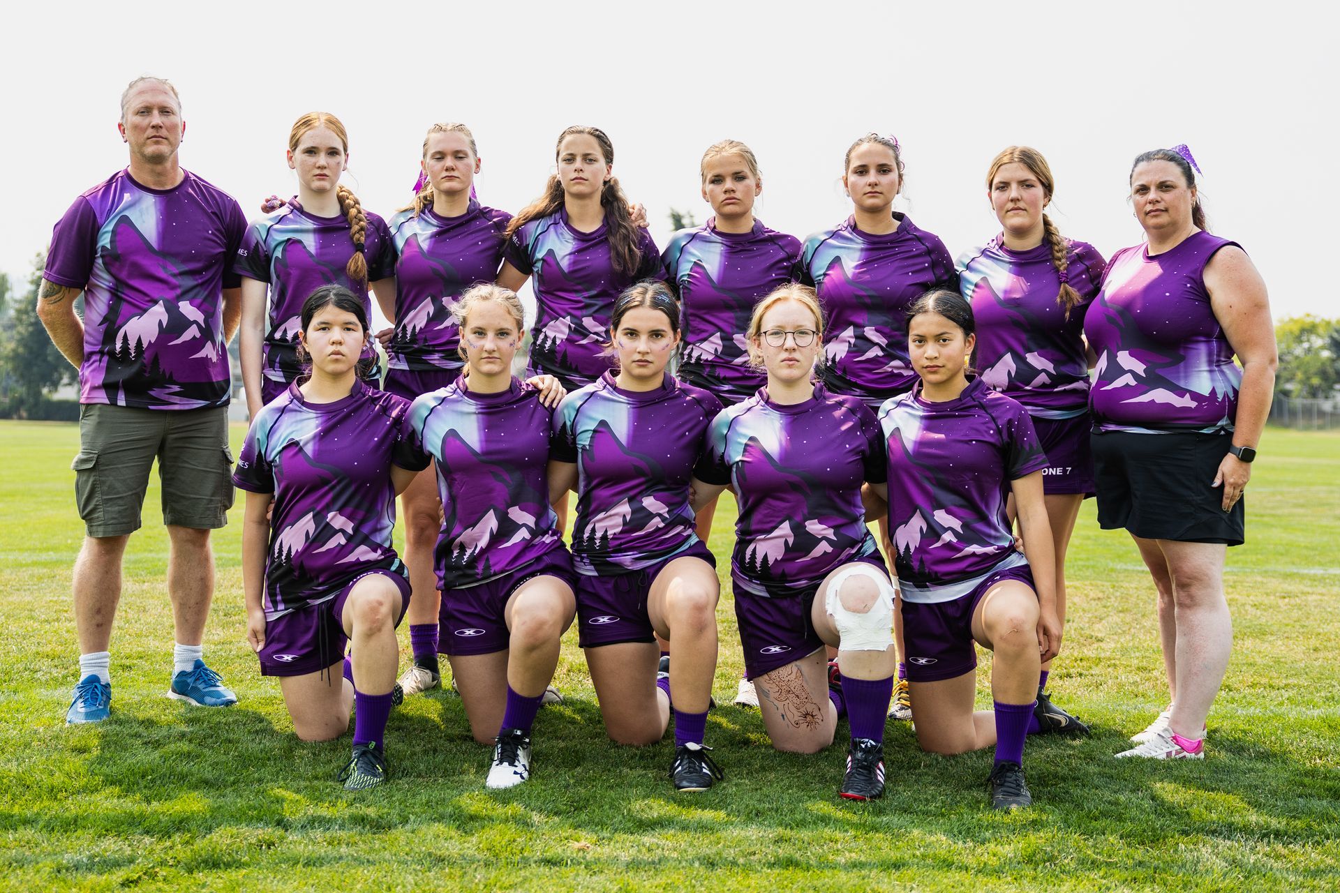 Purple-clad soccer team posing on a grassy field with a coach at left and a ball in front.
