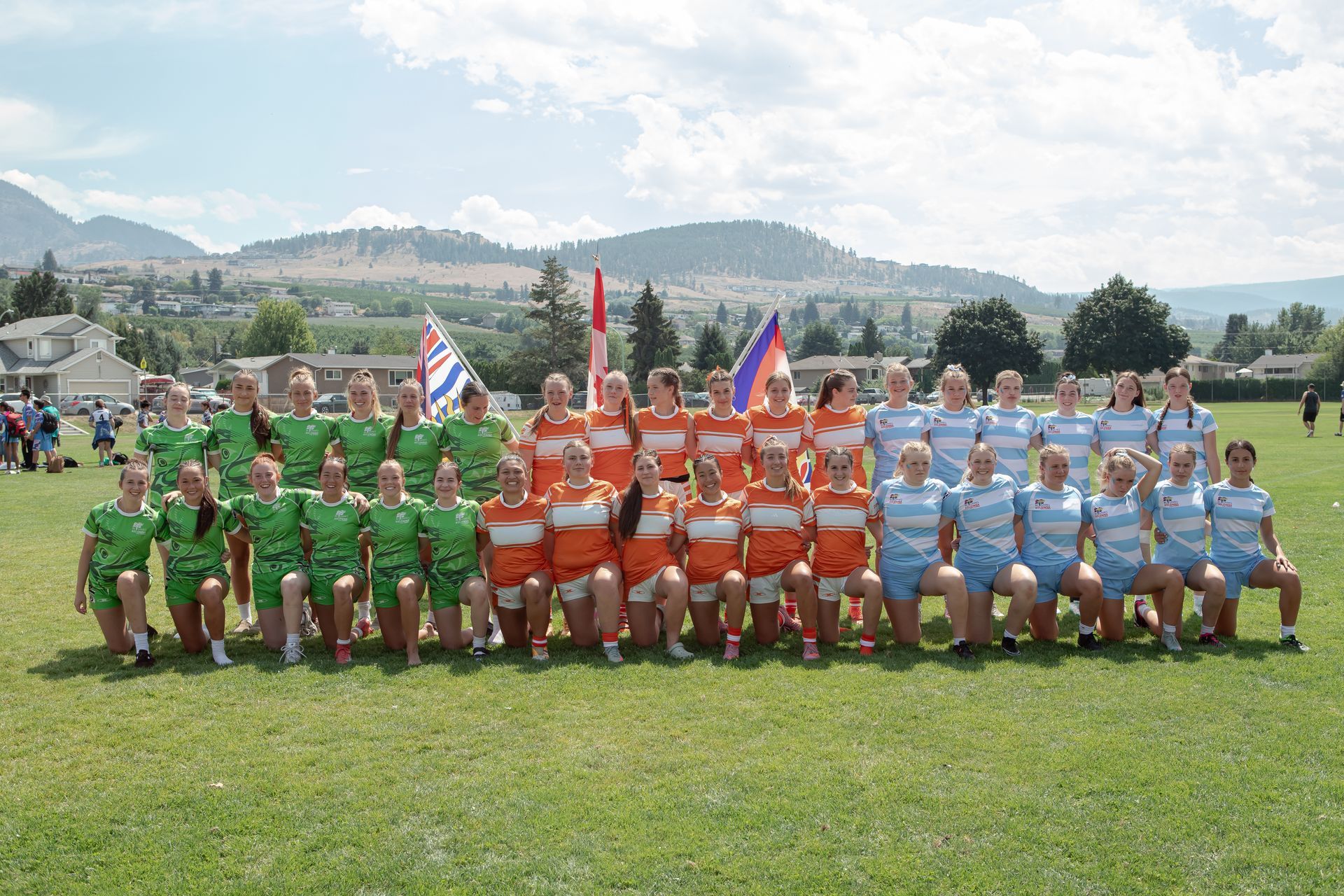 Teams in green, orange, and blue uniforms pose on a grassy field with flags and mountains behind them.