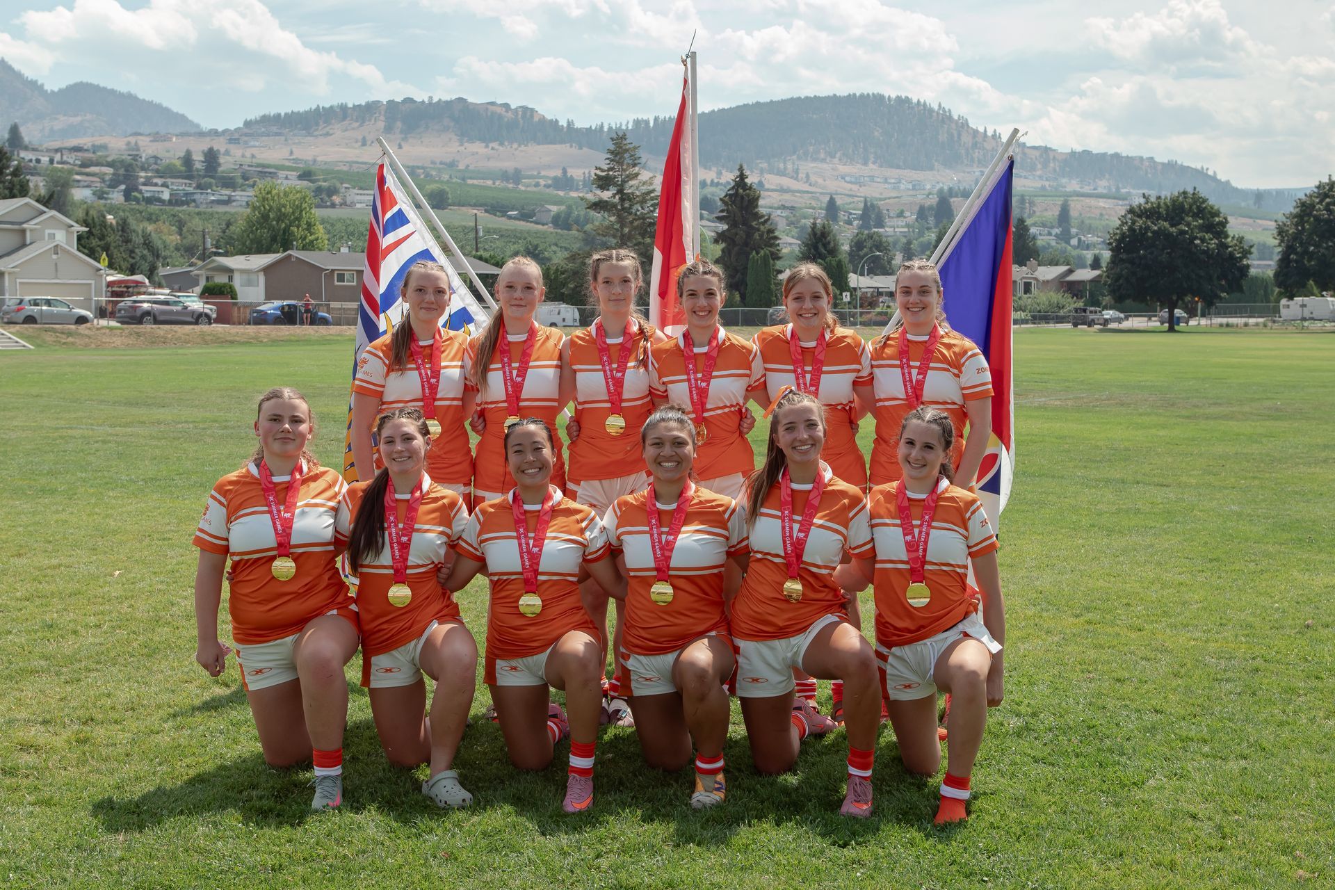 Team of athletes in orange uniforms posing on a grassy field with flags and mountains behind them