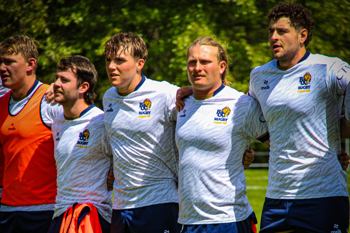 Five soccer players in white jerseys stand arm-in-arm on a field, with one in an orange goalkeeper top
