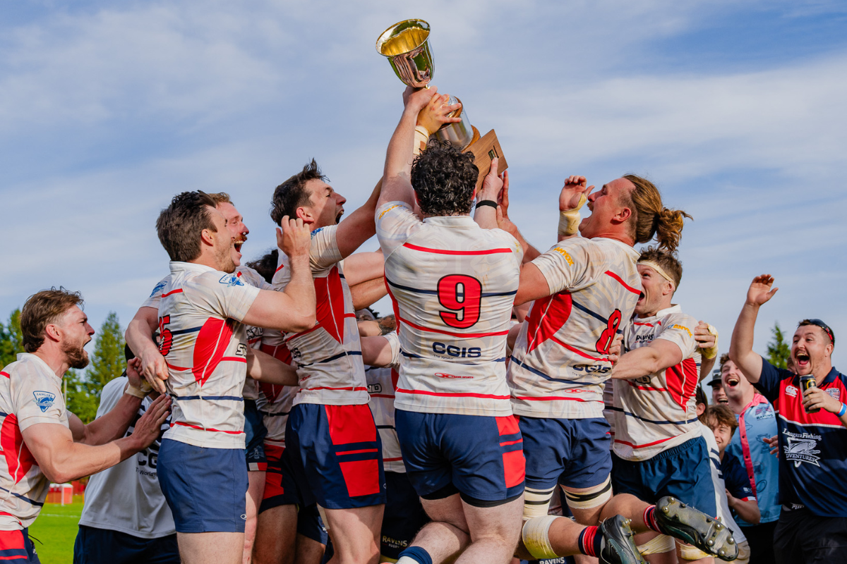 Soccer team in white jerseys celebrates, lifting a gold trophy against a blue sky.