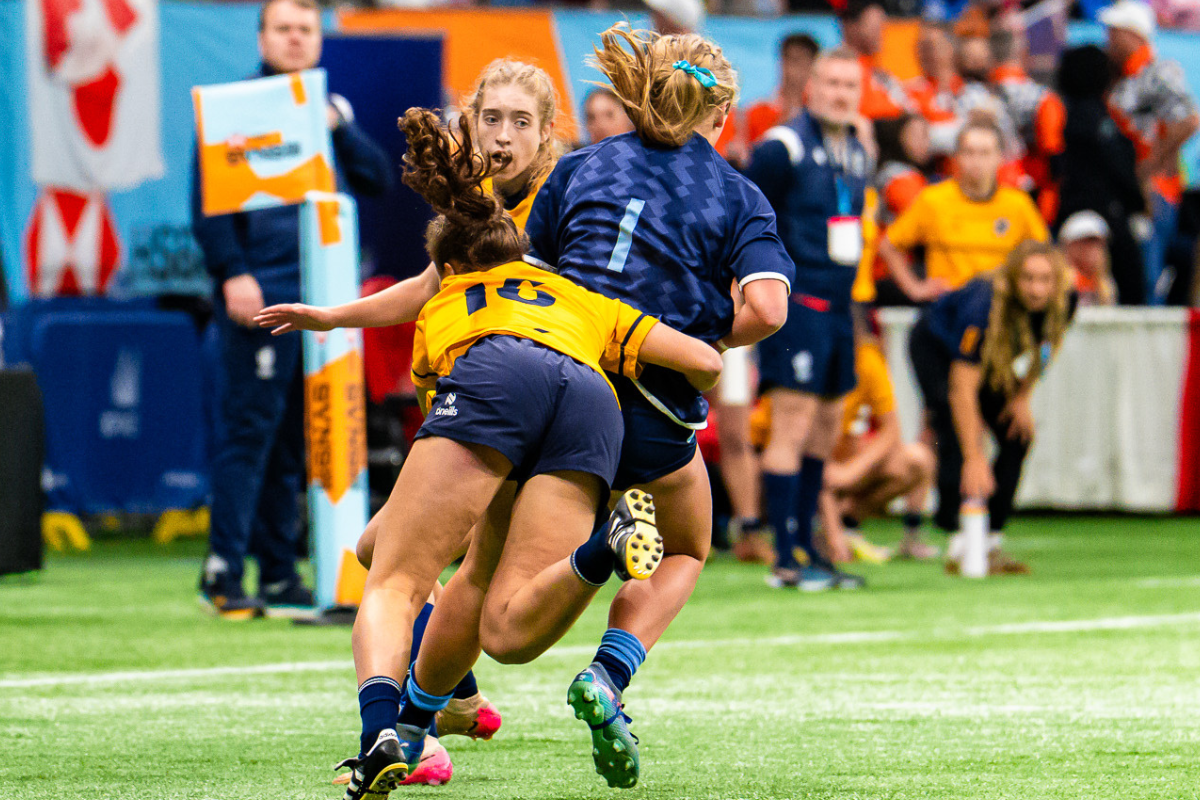 Two rugby players collide on a green field during a match, with spectators and staff in the background.
