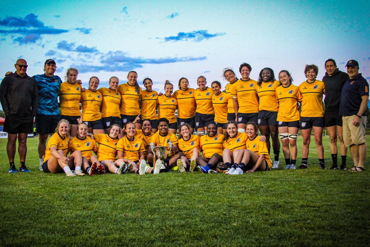 Rugby team posing on a field, wearing yellow jerseys with coaches standing behind them