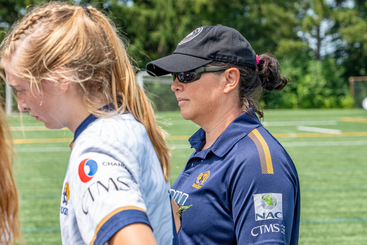 Two women in sports uniforms standing on a grassy field, one wearing a black cap and sunglasses.