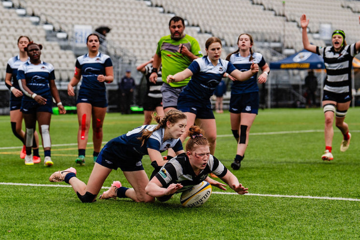 Rugby tackle near the try line, with players in navy and striped kits battling on a wet field