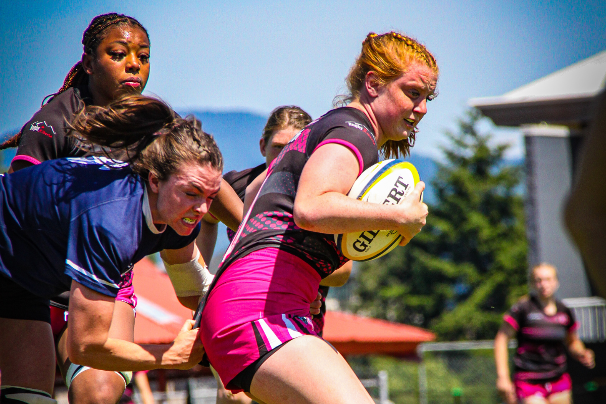 Rugby player in pink sprints with the ball while two defenders tackle her on a sunny field.