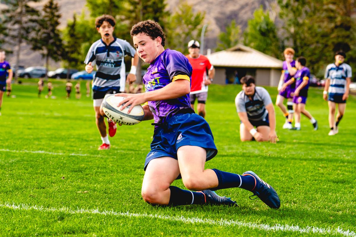 Rugby player in purple runs with the ball on a grassy field while defenders close in behind.