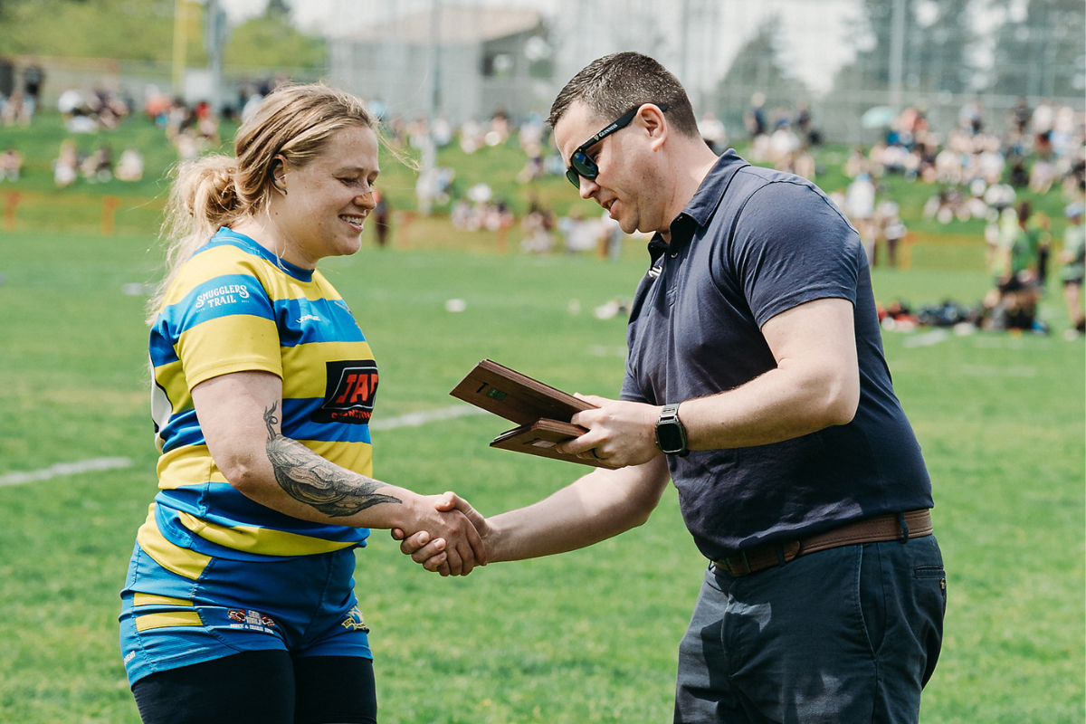 Two people shake hands on a grassy field, one holding a plaque, with a sports crowd in the background.