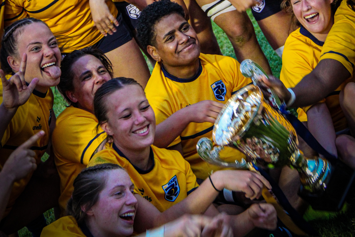 Players in yellow jerseys celebrating and holding up a trophy in a crowded huddle