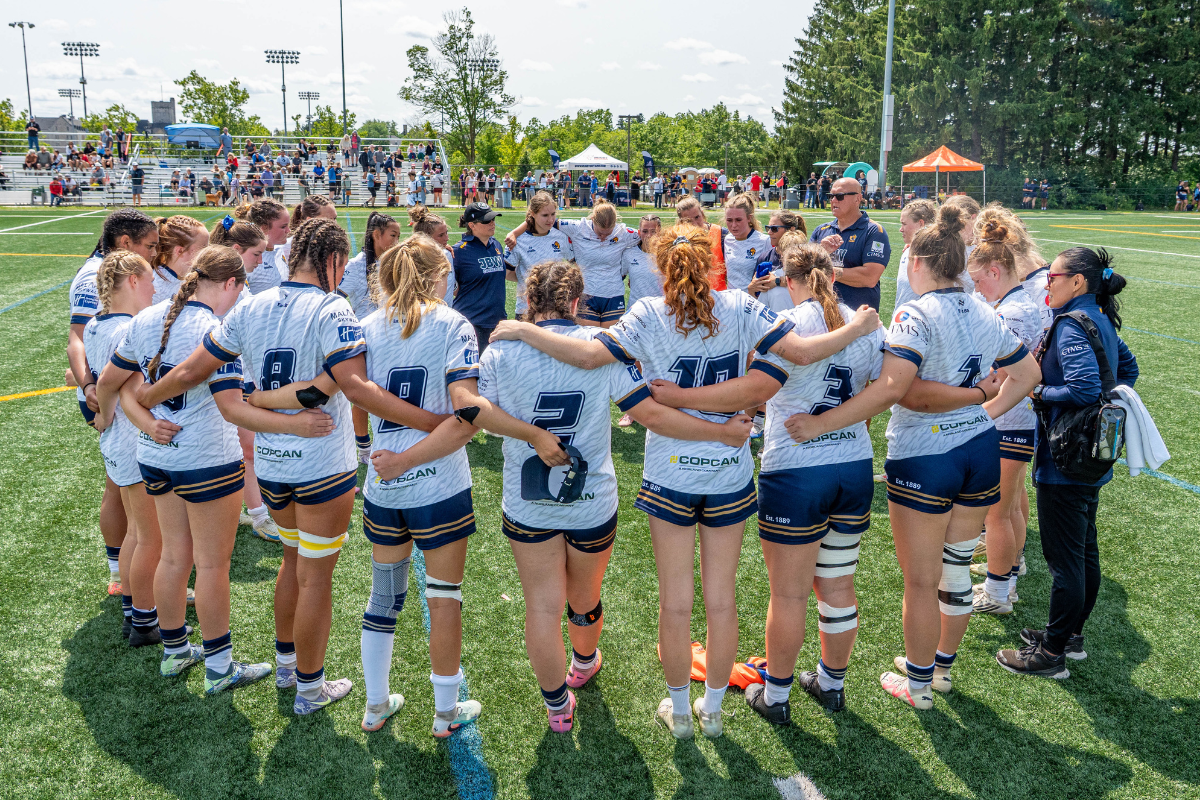 Youth sports team huddled on a grassy field, wearing white jerseys and blue shorts, before a game.