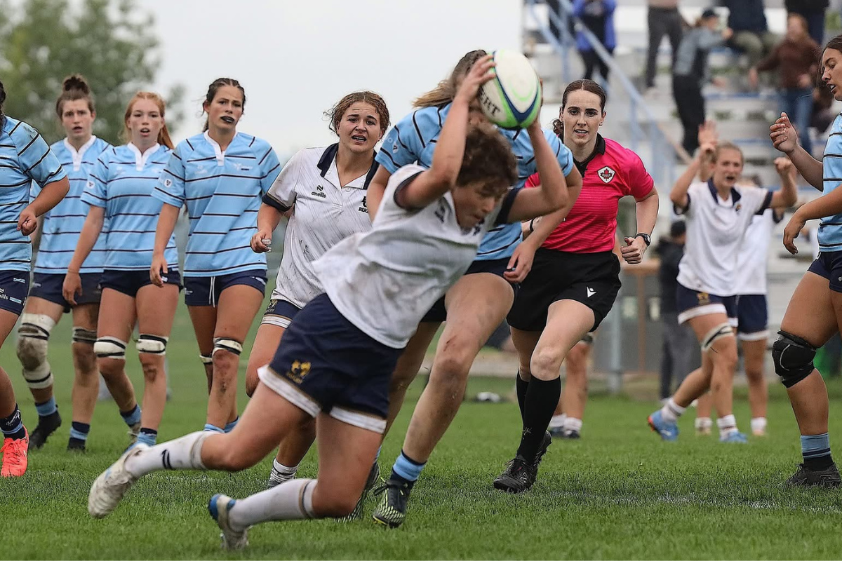 Women playing rugby on a grassy field, one player diving forward while others chase behind.
