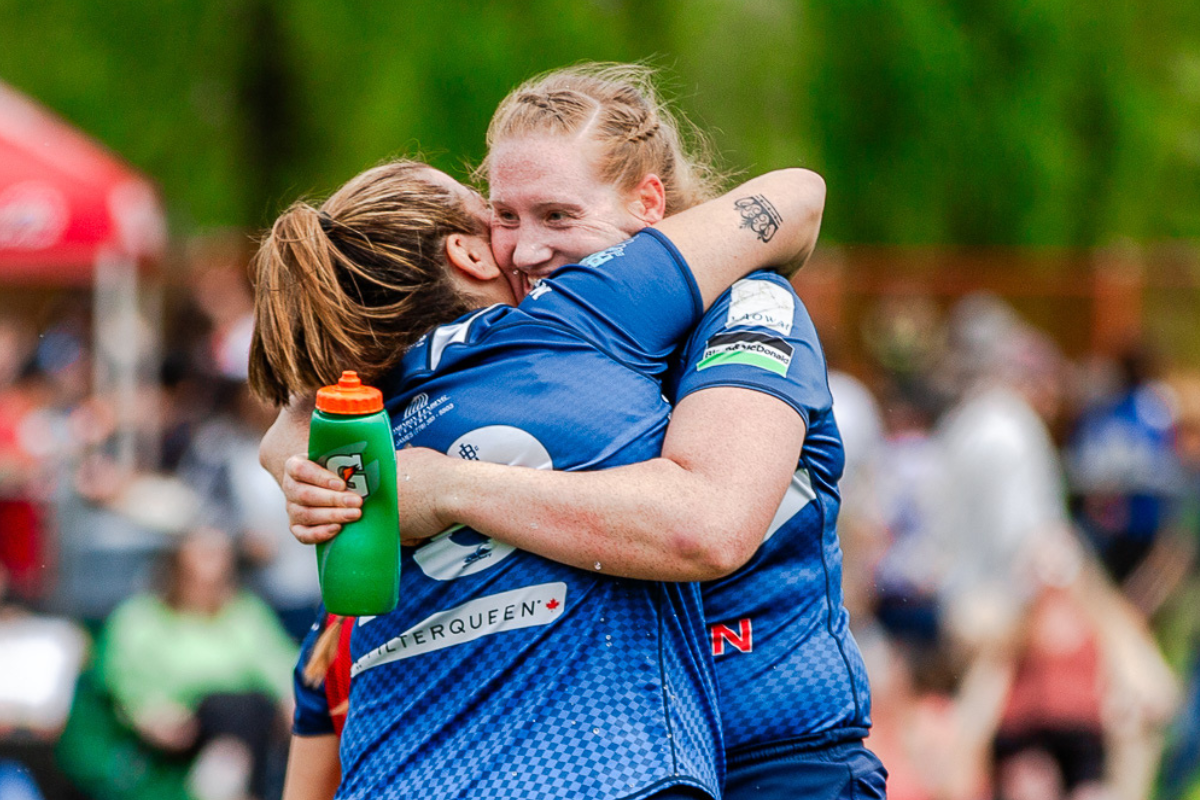 Two athletes in blue jerseys embrace on a sports field, one holding a green water bottle.