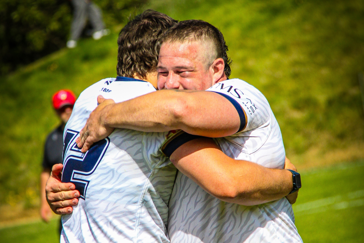 Two rugby players in white jerseys embrace on a grassy field, one smiling with eyes closed.