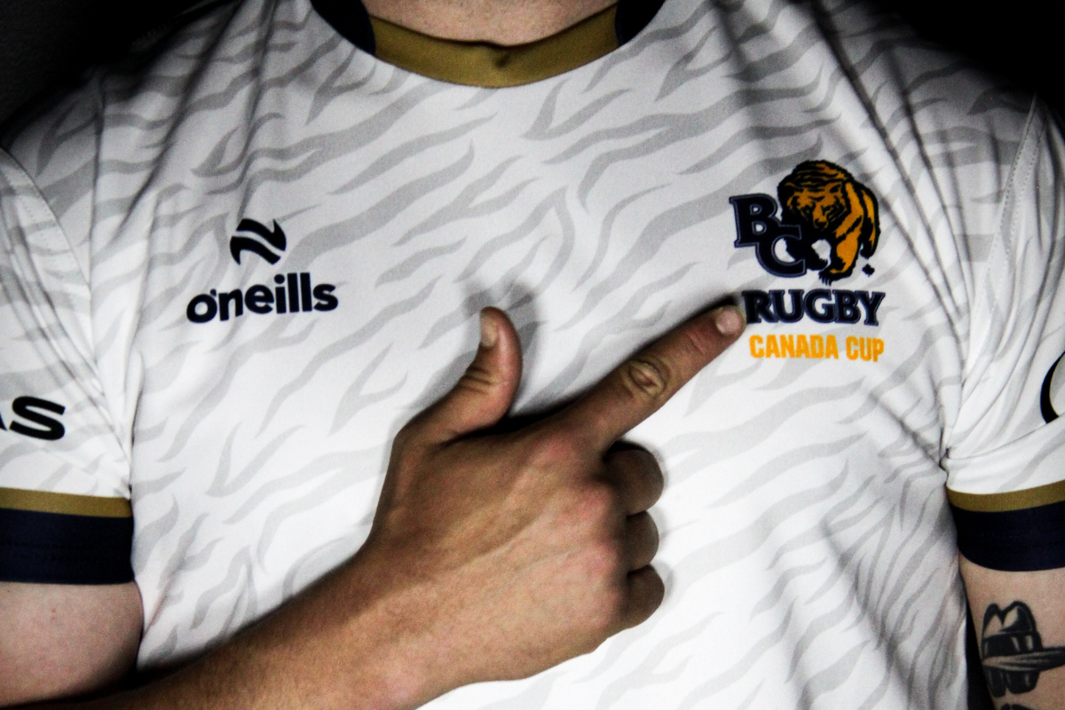 White rugby jersey with tiger logo and “Rugby Canada Cup” text, finger pointing at the chest logo