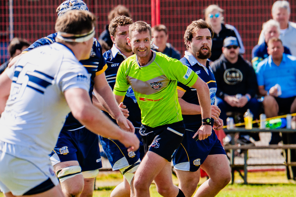 Rugby players in action, with a neon green referee watching closely on the field.