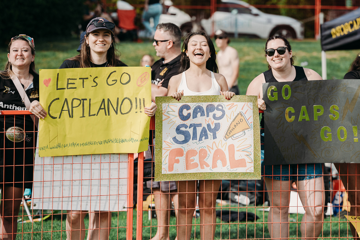 Fans behind a fence hold signs reading “LET’S GO CAPTAINO,” “CAPS STAY FERAL,” and “GO CAPS GO!”