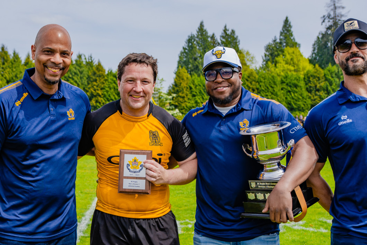 Four men in blue and yellow sports shirts pose outdoors, smiling and holding trophies and a plaque.