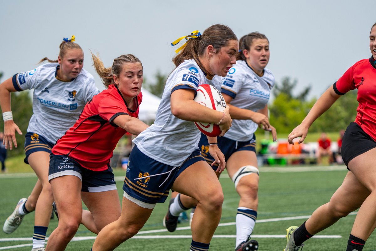 Women rugby players sprinting on a grassy field during a match, with a red-jersey runner in front