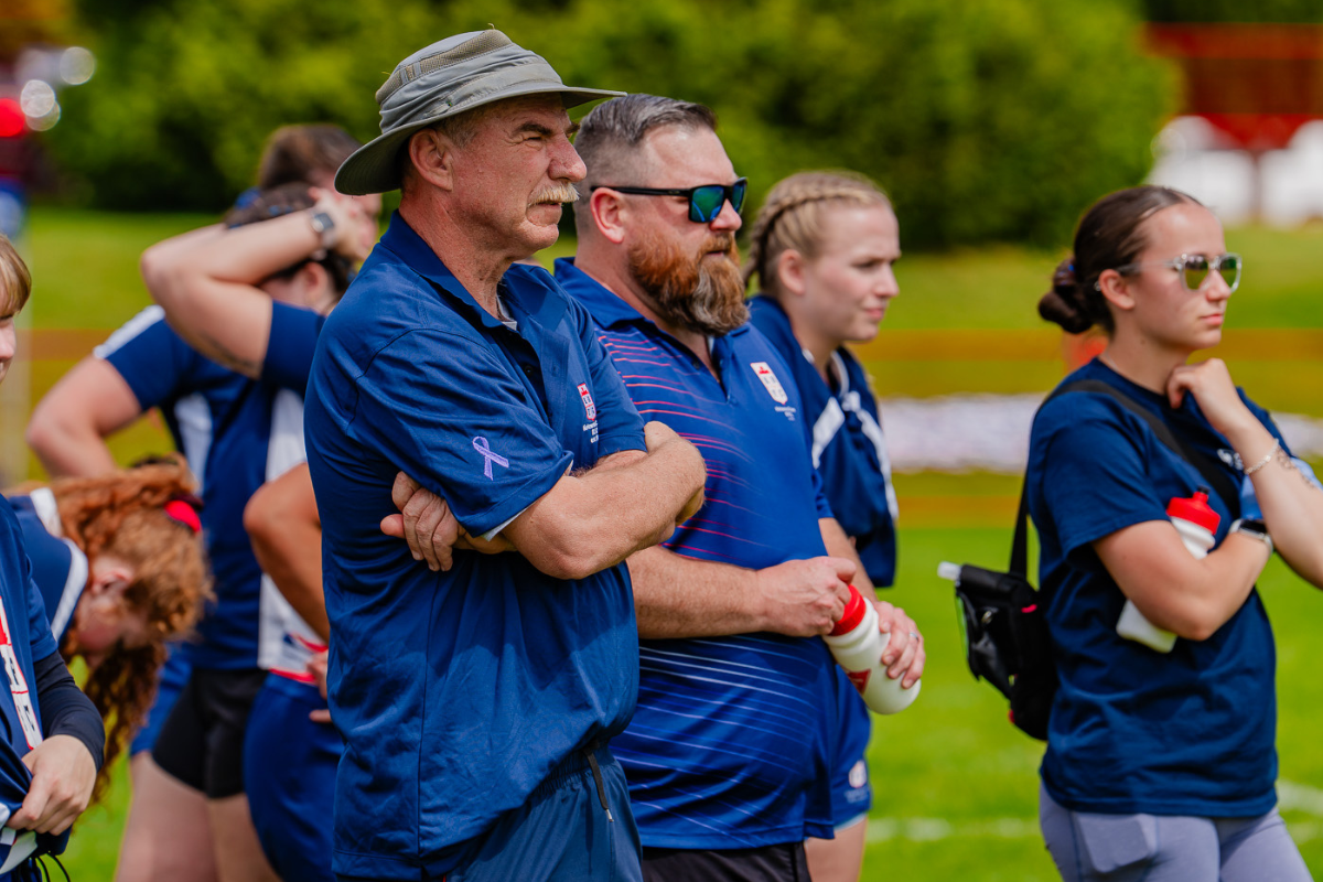 People in blue shirts and hats watching outdoors at a sports event, arms crossed, with green trees behind them