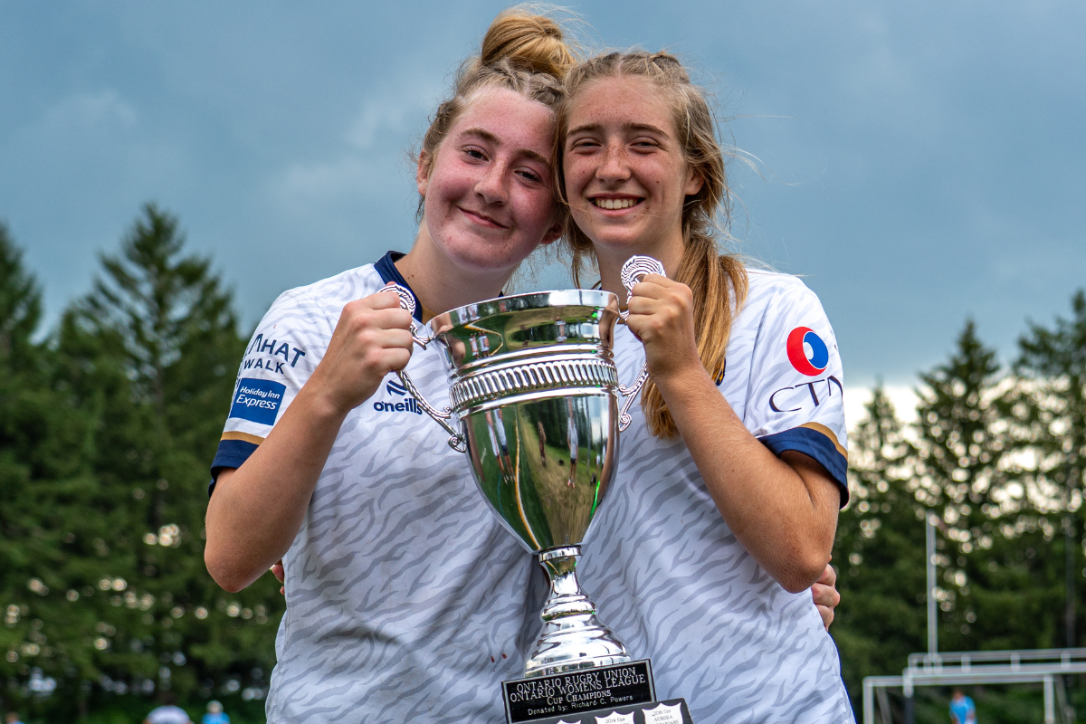 Two athletes in white jerseys hold a large trophy and smile outdoors on a soccer field.
