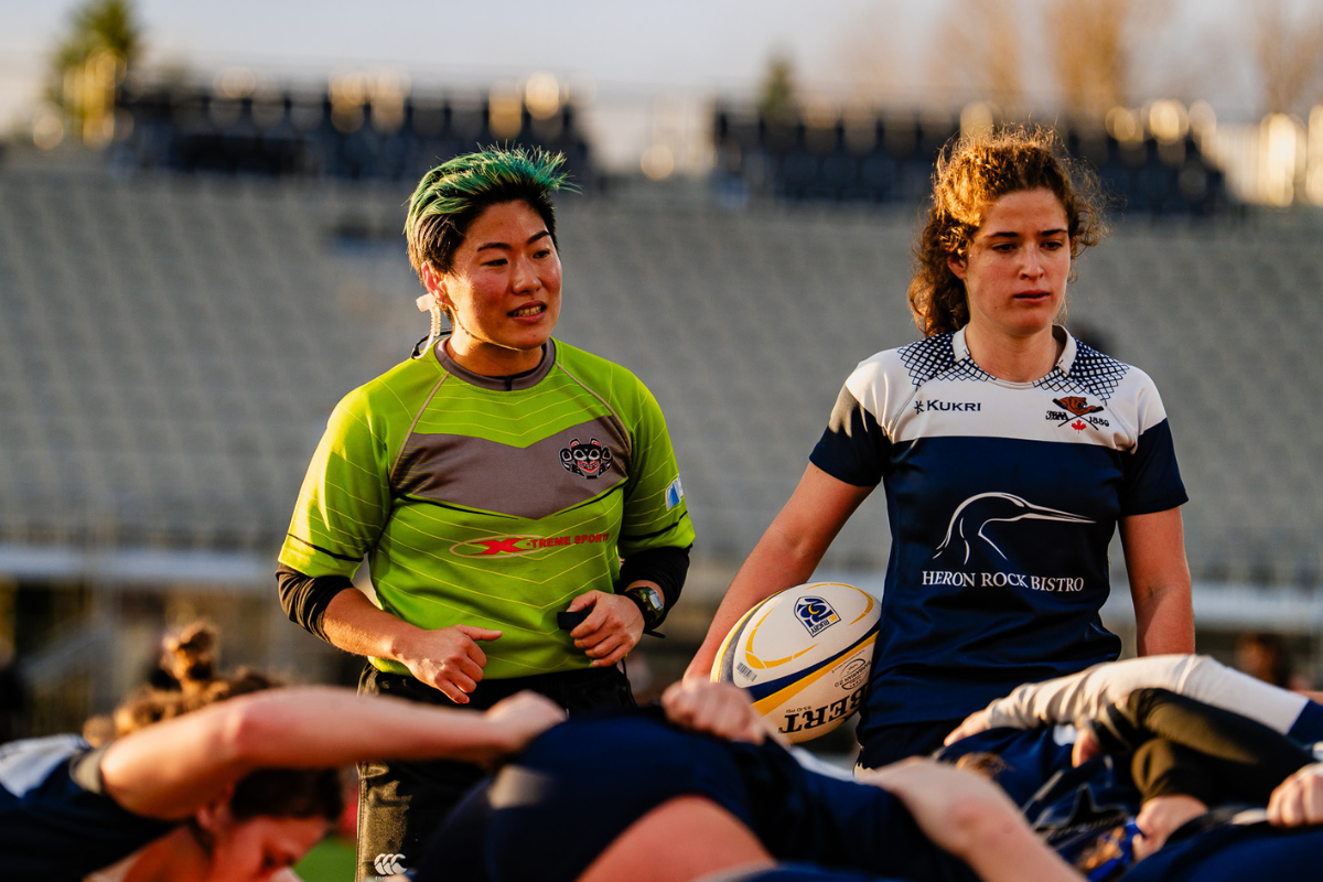 Rugby players huddle on the field, with two athletes in green and navy jerseys in front.