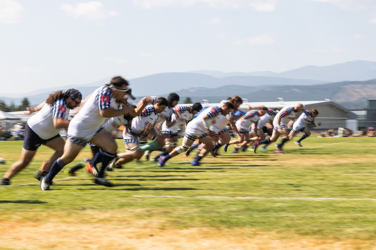 Rugby players sprint across a grassy field during a match, with mountains in the background.