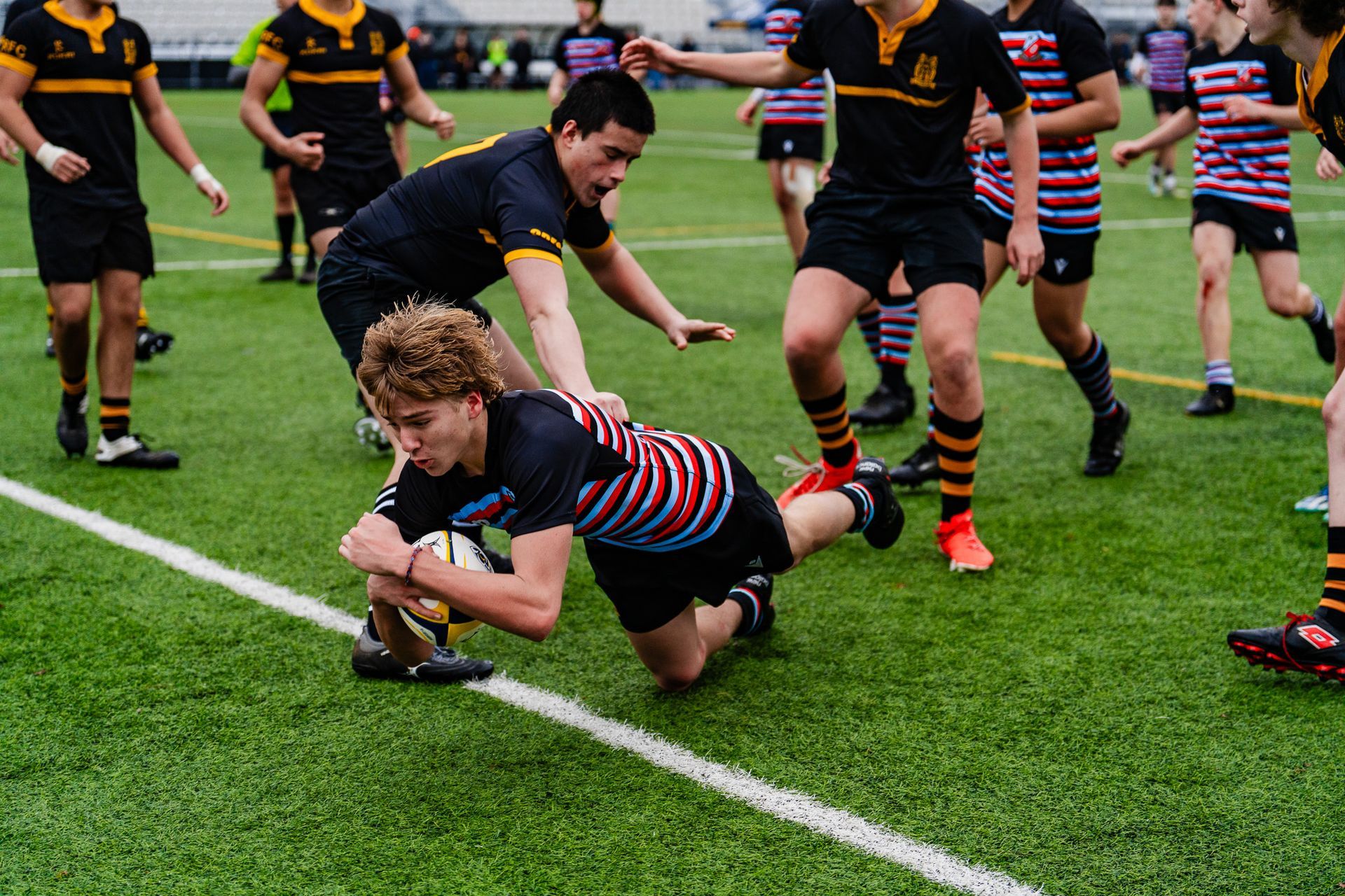 Rugby player in striped jersey tackled on green field, holding the ball near the sideline.