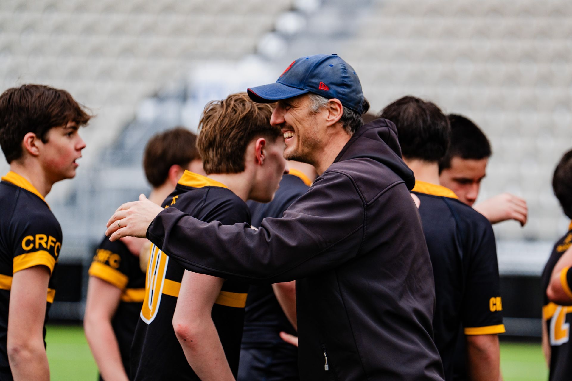 Coach hugging a player on a sports field, with teammates in black and yellow jerseys nearby