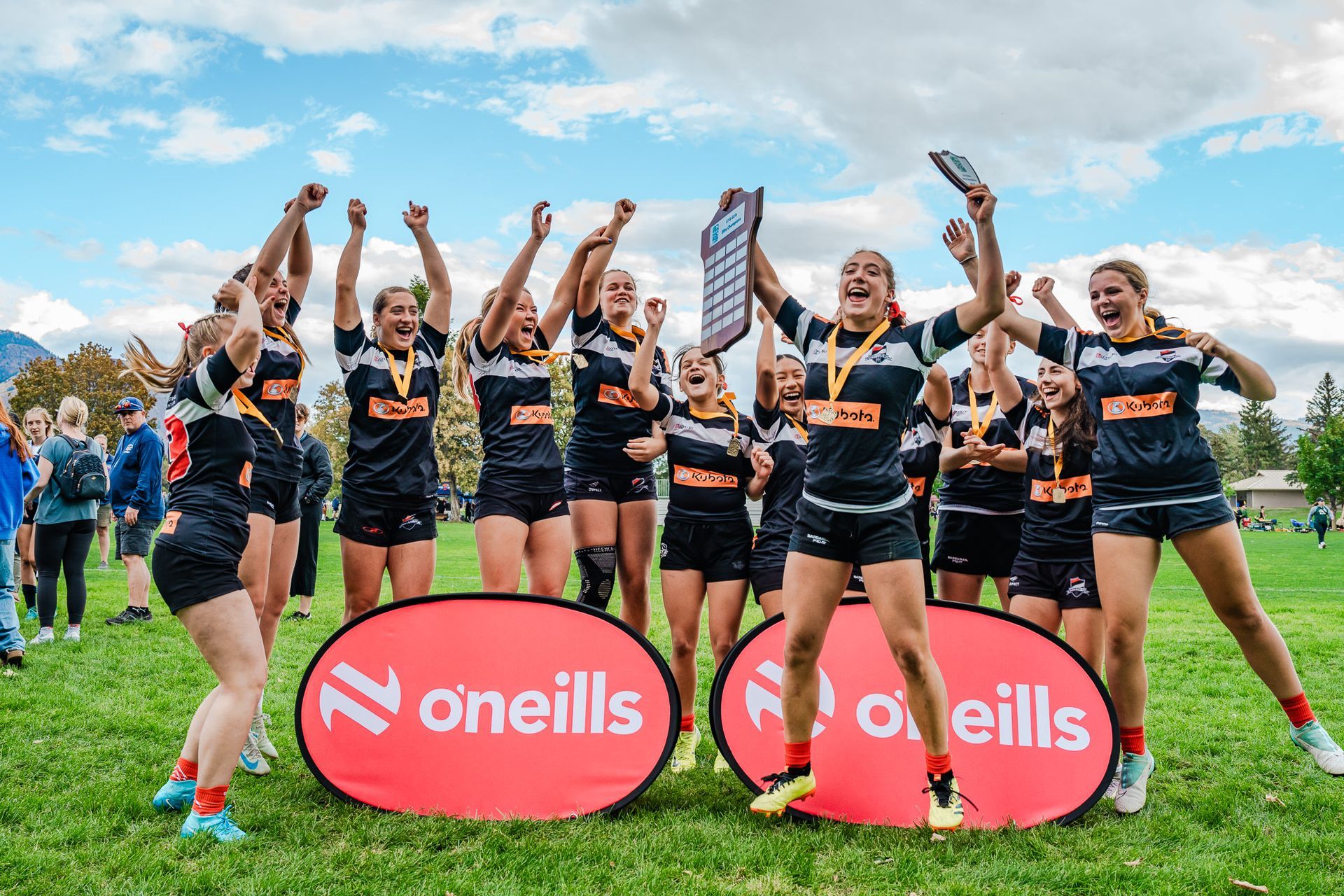 Women’s sports team celebrating with raised arms on a grassy field beside O’Neills banners