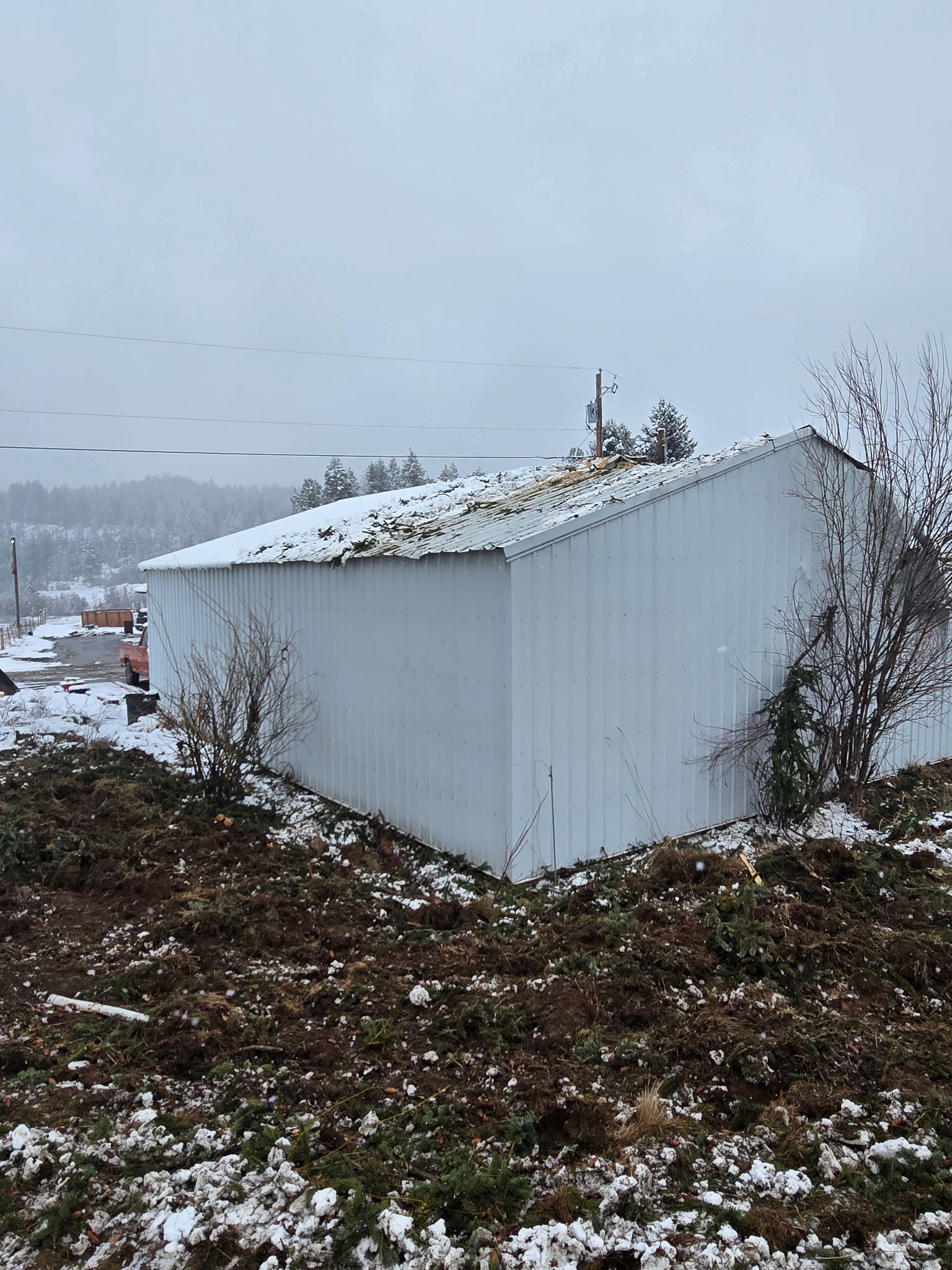 A white metal shed sits on snowy ground in an overcast outdoor landscape with bare trees and a distant hillside.