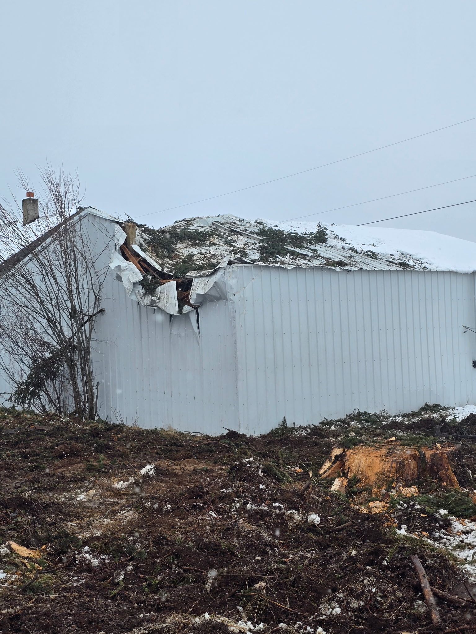 A white building with a damaged roof and wall, surrounded by dark, muddy ground with patches of snow.