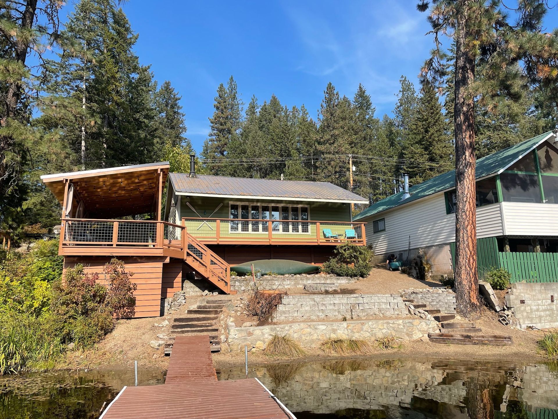 Green cabin with a wooden deck and staircase leading down to a waterfront dock, set against a forest backdrop.