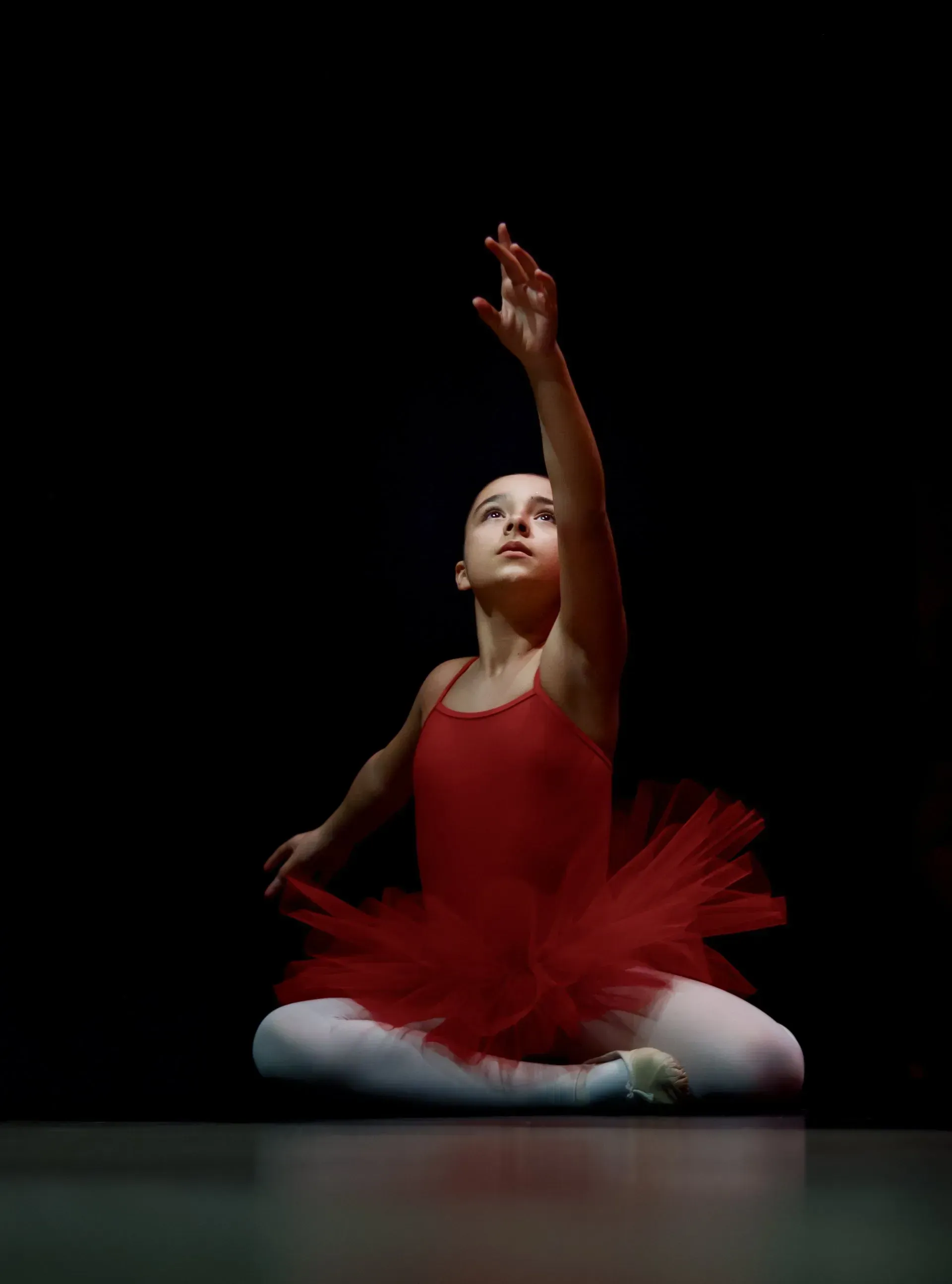 Young ballet dancer in a red tutu poses on stage, arm raised against a dark background.